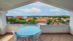 A balcony with a checkered table and chairs, overlooking a coastal town with terracotta-roofed buildings and a clear sky.