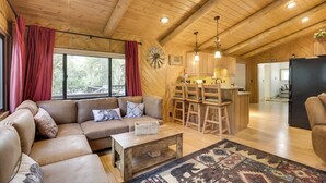 Interior acogedor de una cabaña con techo de madera, una sala de estar con sofá y mesa de centro, y una cocina con armarios de madera y refrigerador.