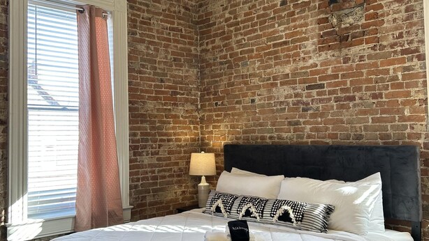 Een slaapkamer met een bakstenen muur, een bed met witte beddengoed, een plafondventilator en een raam met jaloezieën.