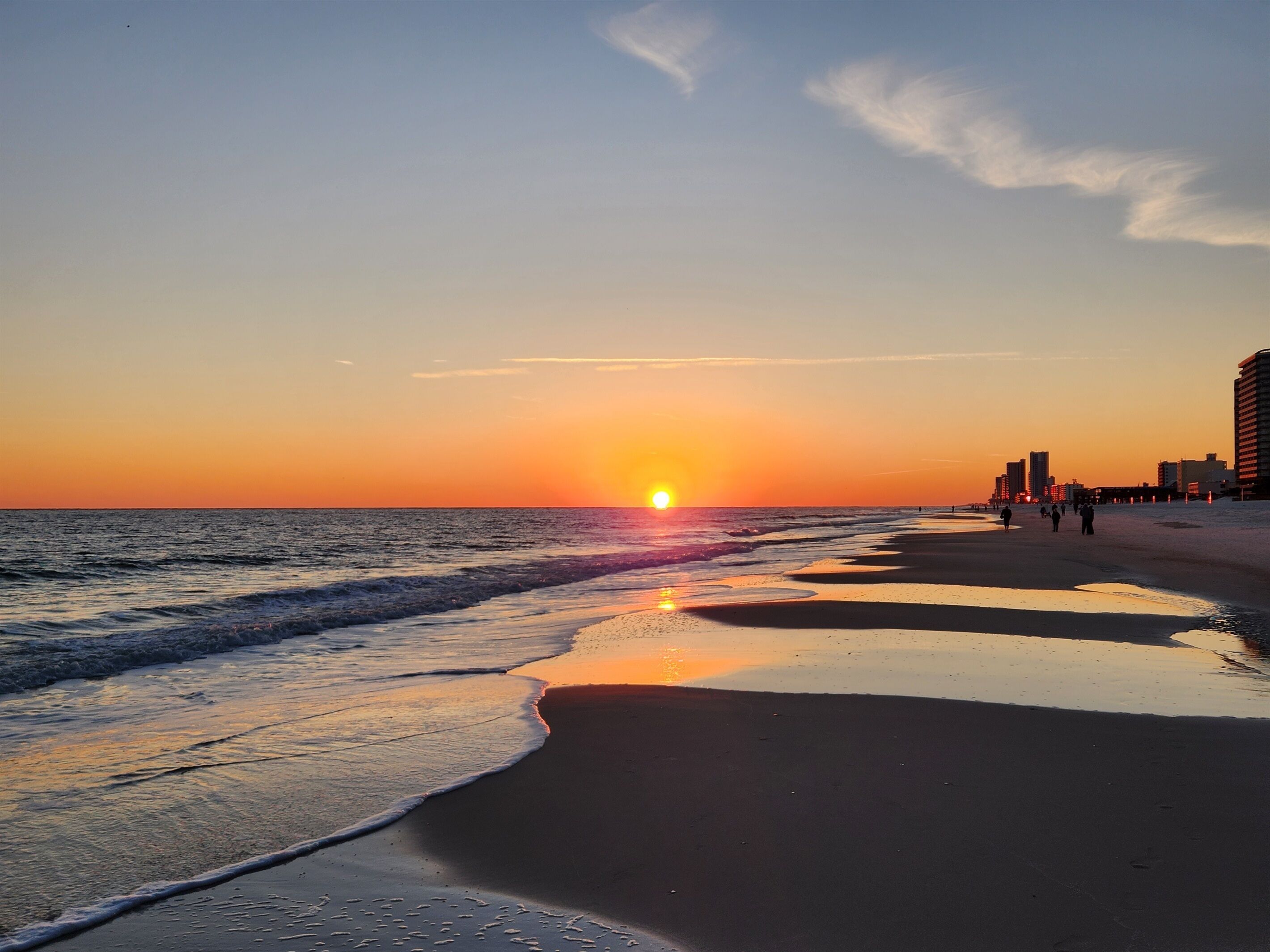 A beach with the sun setting over the horizon, waves gently lapping the shore, and a pier extending into the water.