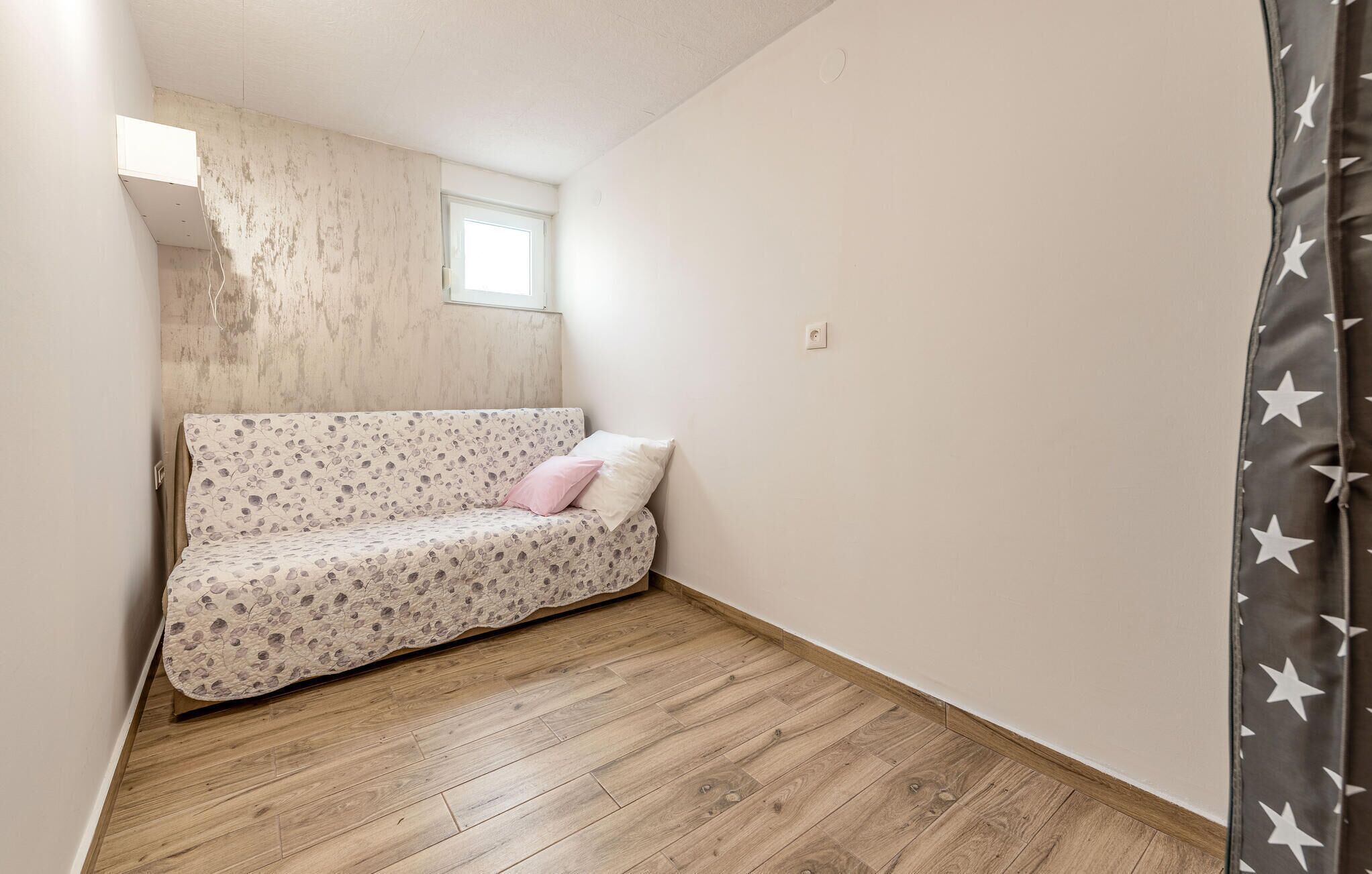 Habitació petita i estreta amb terra de fusta, un llit sol amb col·cha amb motius florals i una finestra que deixa entrar llum natural.