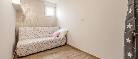 Habitació petita i estreta amb terra de fusta, un llit sol amb col·cha amb motius florals i una finestra que deixa entrar llum natural.