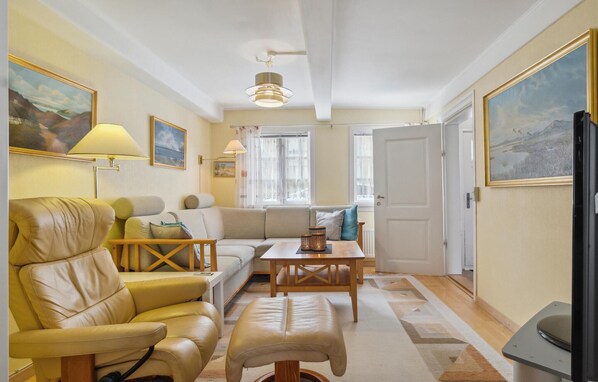Un salón acogedor con un sofá, un sillón y un reposapiés, una mesa de centro de madera, y una mesa de comedor. Hay cuadros en las paredes, una lámpara en el techo y una puerta que da a otra habitación.