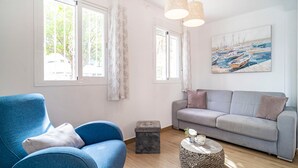 Una sala de estar con un sillón azul, un sofá gris, un pequeño reposapiés y una mesa de centro redonda. Hay dos ventanas con cortinas, un cuadro de barcos en la pared y una lámpara colgante.