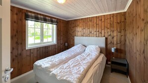 Un dormitorio con pared de paneles de madera, un ventanal con vistas, una cama con ropa de cama blanca y una lámpara de noche.