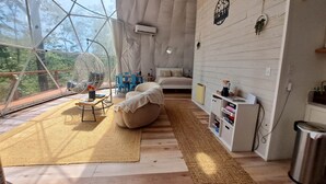 Una habitación amplia con un gran ventanal abovedado, un sillón colgante, una cama, una mesita y una estantería.