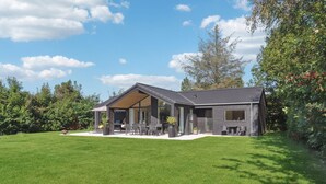 Et moderne hus med en overdækket terrasse, græsplæne og omgivende træer.
