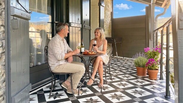 Un espace extérieur abrité, aménagé avec une terrasse carrelée, une table sur laquelle se trouve une assiette de nourriture et des verres à vin, ainsi que des plantes en pot.