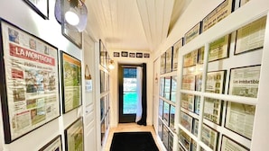 Un couloir dont les murs sont ornés de coupures de journaux encadrées, un tapis noir sur le sol et une porte donnant sur un espace extérieur.