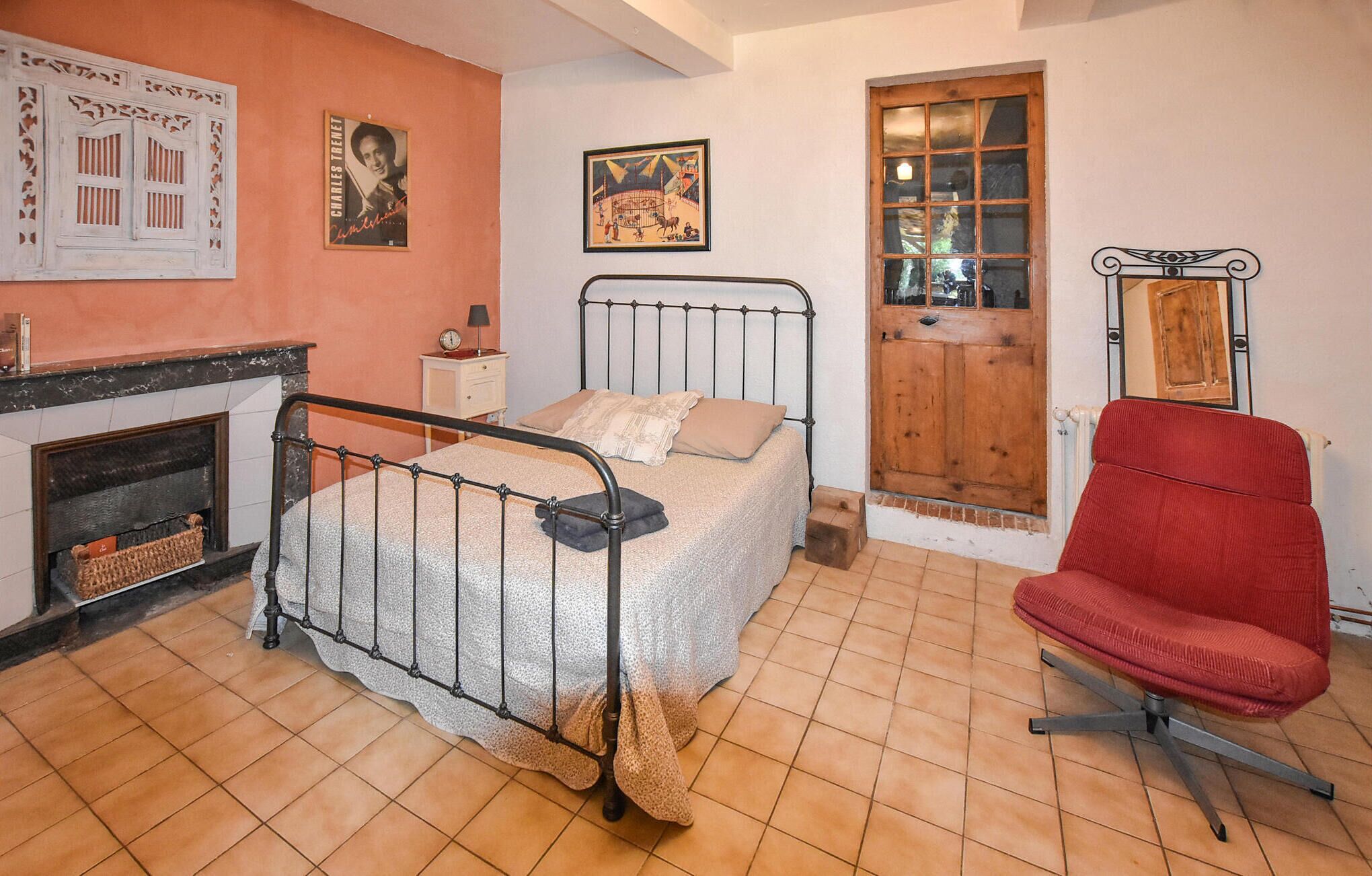 Une chambre avec un lit en métal, une chaise rouge, une porte en bois et une cheminée.