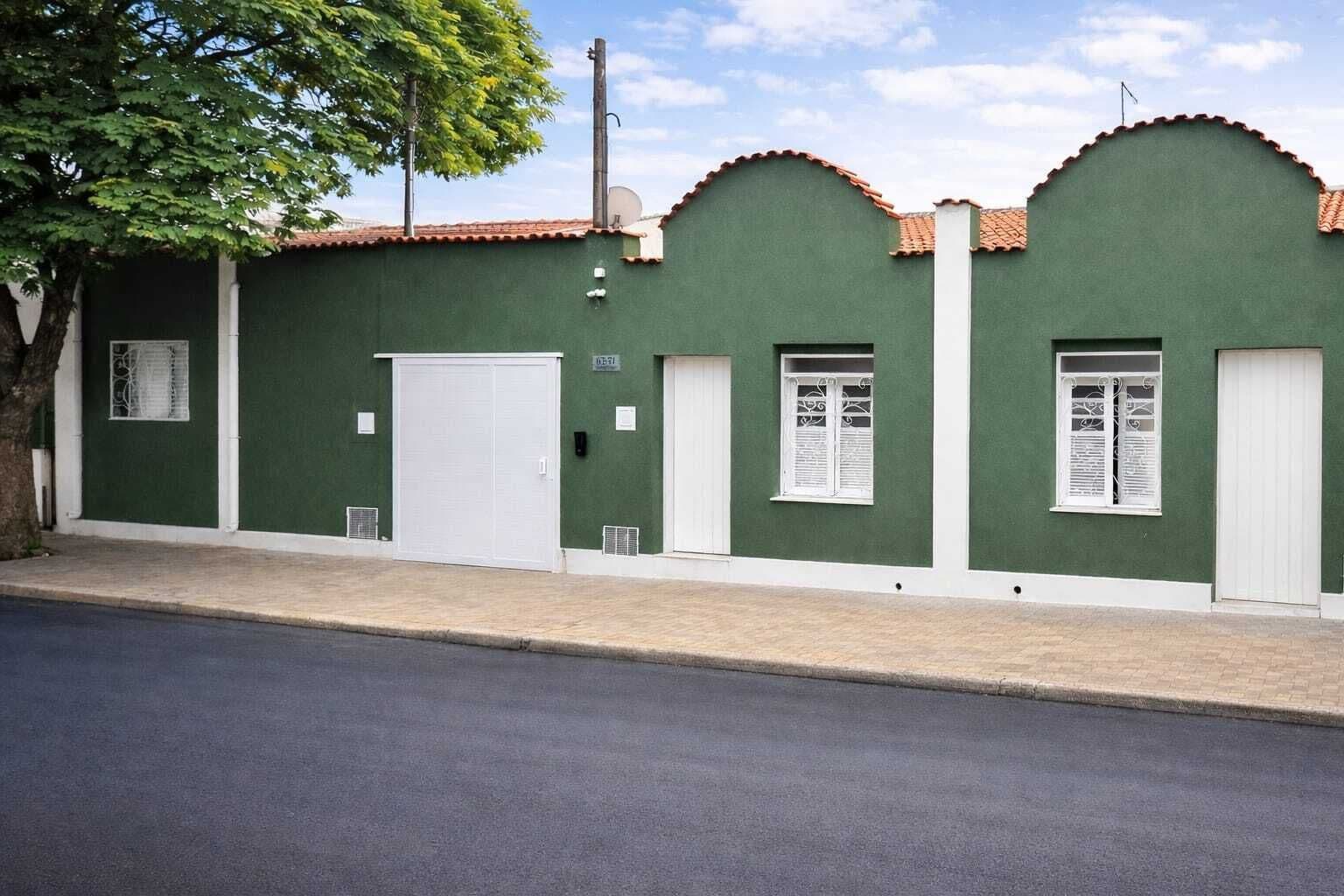 Um edifício verde com uma porta branca, duas janelas com contraportas brancas e uma árvore em frente.