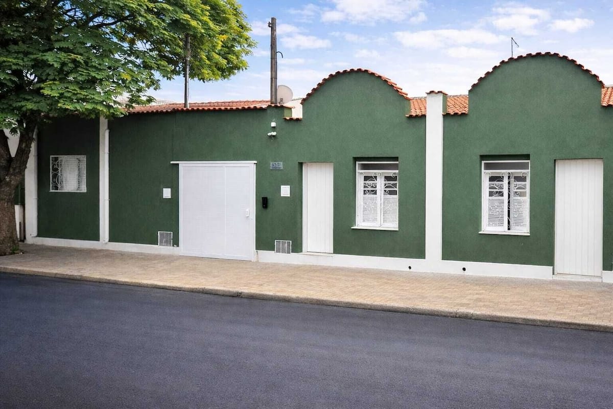 Un edificio verde con una puerta blanca, dos ventanas con contraventanas blancas y un árbol en frente.