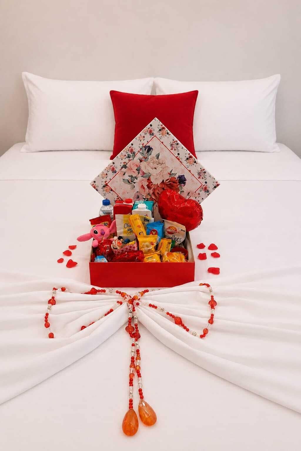 Una cama bien tendida con una almohada roja, una caja de bombones estampada con motivos florales y un globo con forma de corazón.