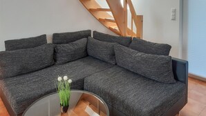 Un sofá modular con tela texturizada, una mesa de centro de vidrio con una planta en maceta y una escalera de madera al fondo.