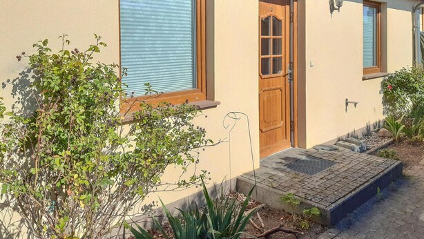 Una casa con puerta de madera, una ventana con persianas y un pequeño jard ín con plantas y entrada pavimentada.