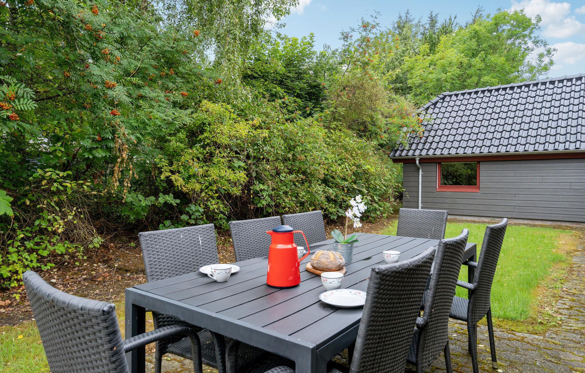 Ein Essbereich im Freien mit Tisch und Stühlen, einem roten Wasserkocher, einem Brot und einem kleinen Haus im Hintergrund.