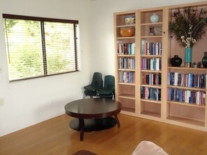 Ein Zimmer mit einem runden Holztisch, zwei grünen Stühlen, einem Bücherregal voller Bücher und einem Fenster mit Jalousien.