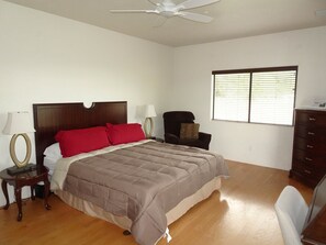 Une chambre à coucher comprenant un lit, deux tables de chevet, une chaise, une commode, un ventilateur de plafond et une fenêtre avec des stores.