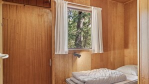 A small wooden cabin room with a single bed, a window with curtains, and a view of trees outside.