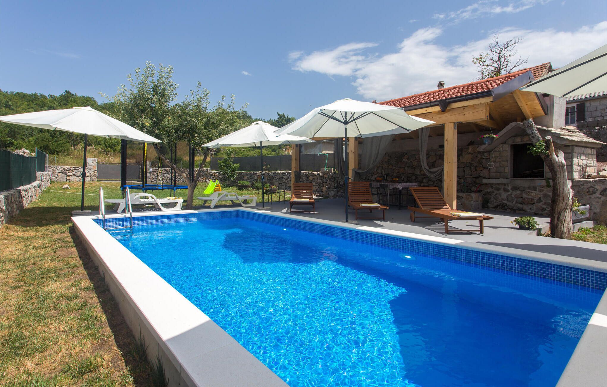 Una piscina rectangular amb aigua blava cristal·lina, envoltada per una paret de pedra i un pont de fusta. Hi ha diversos para-sols blancs que fan ombra, i unes quantes gandules col·locades al voltant del pont.