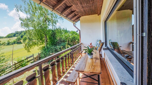 A balcony with a wooden table and benches, overlooking a green landscape.