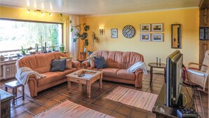 A cozy living room with a brown leather sofa, a wooden coffee table, and a television. There are potted plants, a clock, and framed pictures on the walls.