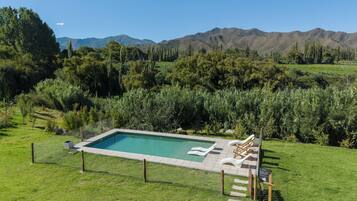 Una piscina rectangular con superficie de agua azul cristalina, rodeada por un césped con una terraza de madera y sillones de descanso.