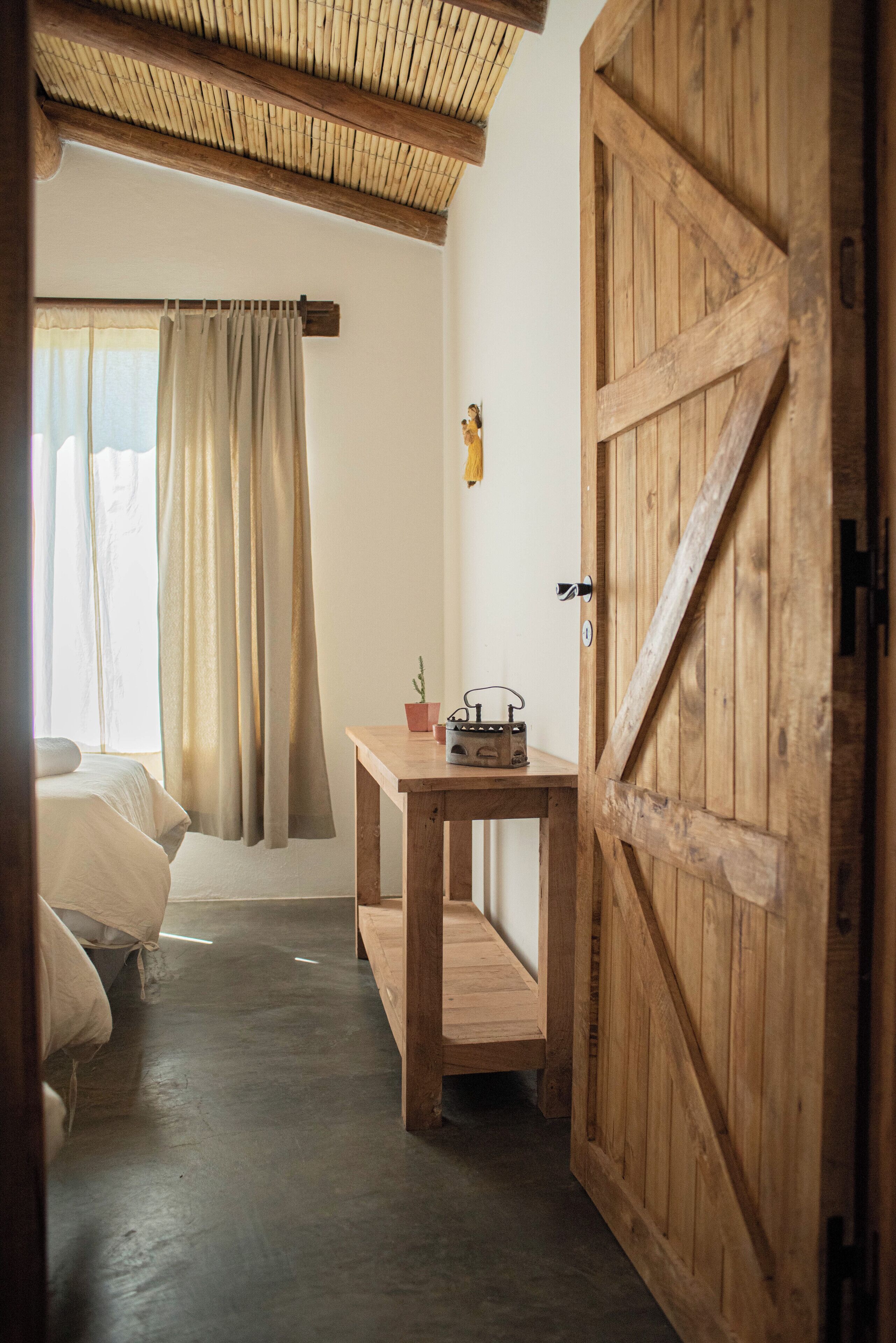 A room with a wooden door, a bed, a table with a radio, and a window with curtains.