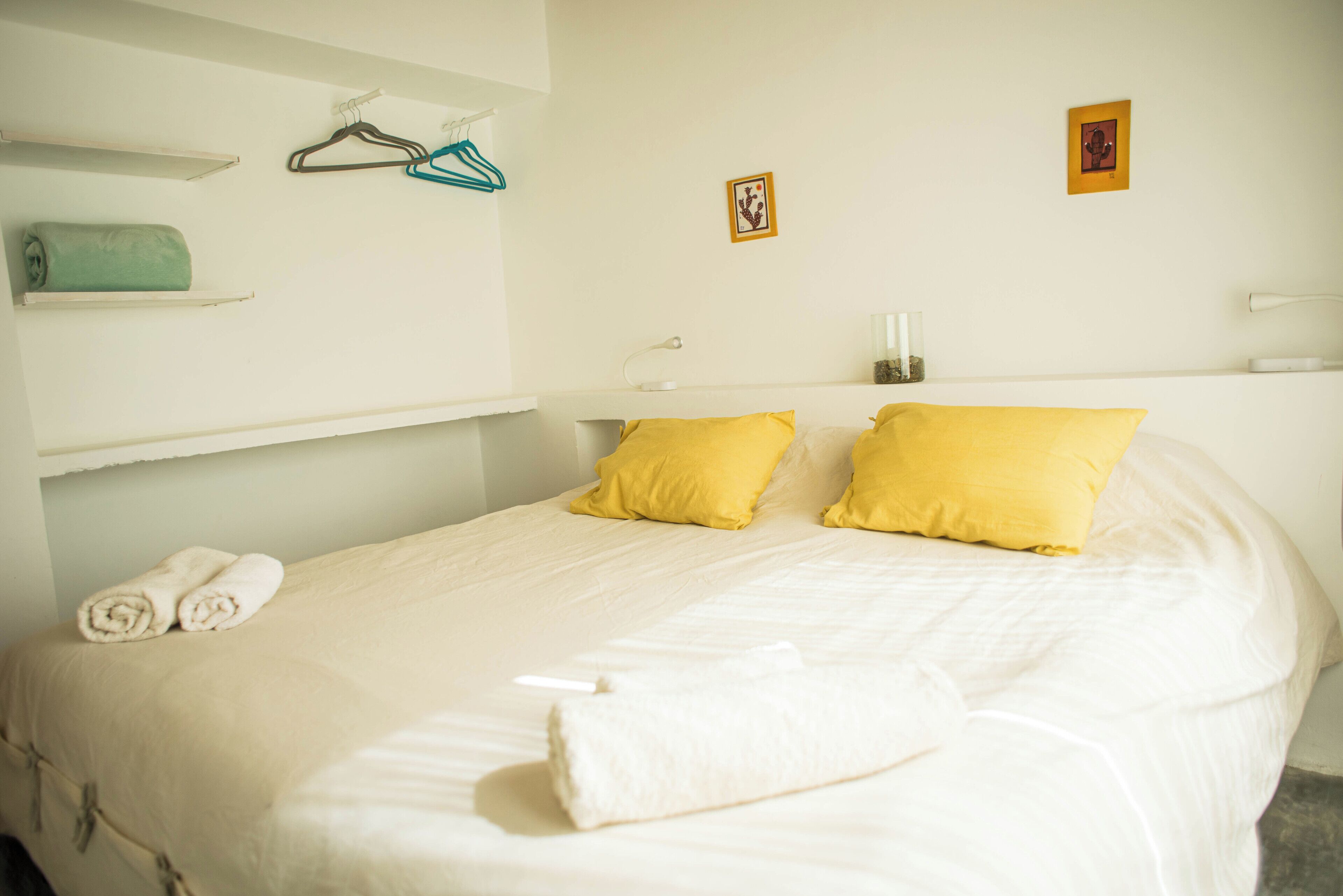 Cama bem arrumada com almofadas brancas e amarelas, uma toalha enrolada e uma pequena estante com cabides.