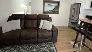 A living room with a brown sofa, a patterned rug, a painting of a mountain landscape, a kitchen with a refrigerator, and a dining area with a bar stool. - Modern studio in MarysVille – A Forest Haven (Marysville)