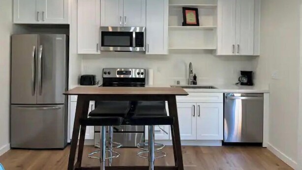 A modern kitchen with white cabinets, stainless steel appliances, a wooden table, and bar stools. - Modern studio in MarysVille – A Forest Haven (Marysville)