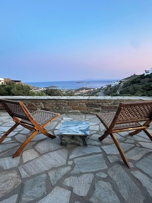 A stone patio with two wicker chairs and a marble table, overlooking a coastal landscape with buildings and a clear sky.