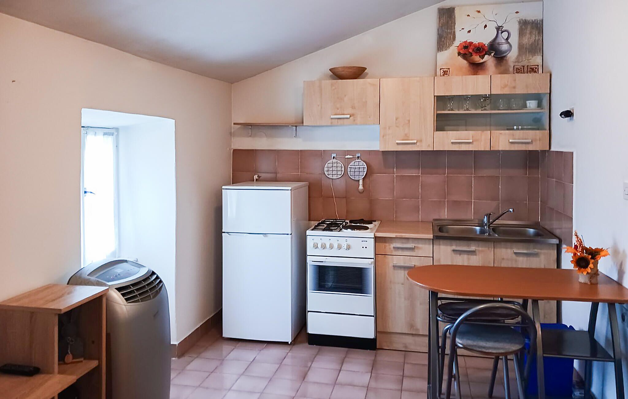 Een kleine keuken met een koelkast, fornuis en spoelbak. Er is een eethoek met een tafel en stoelen.