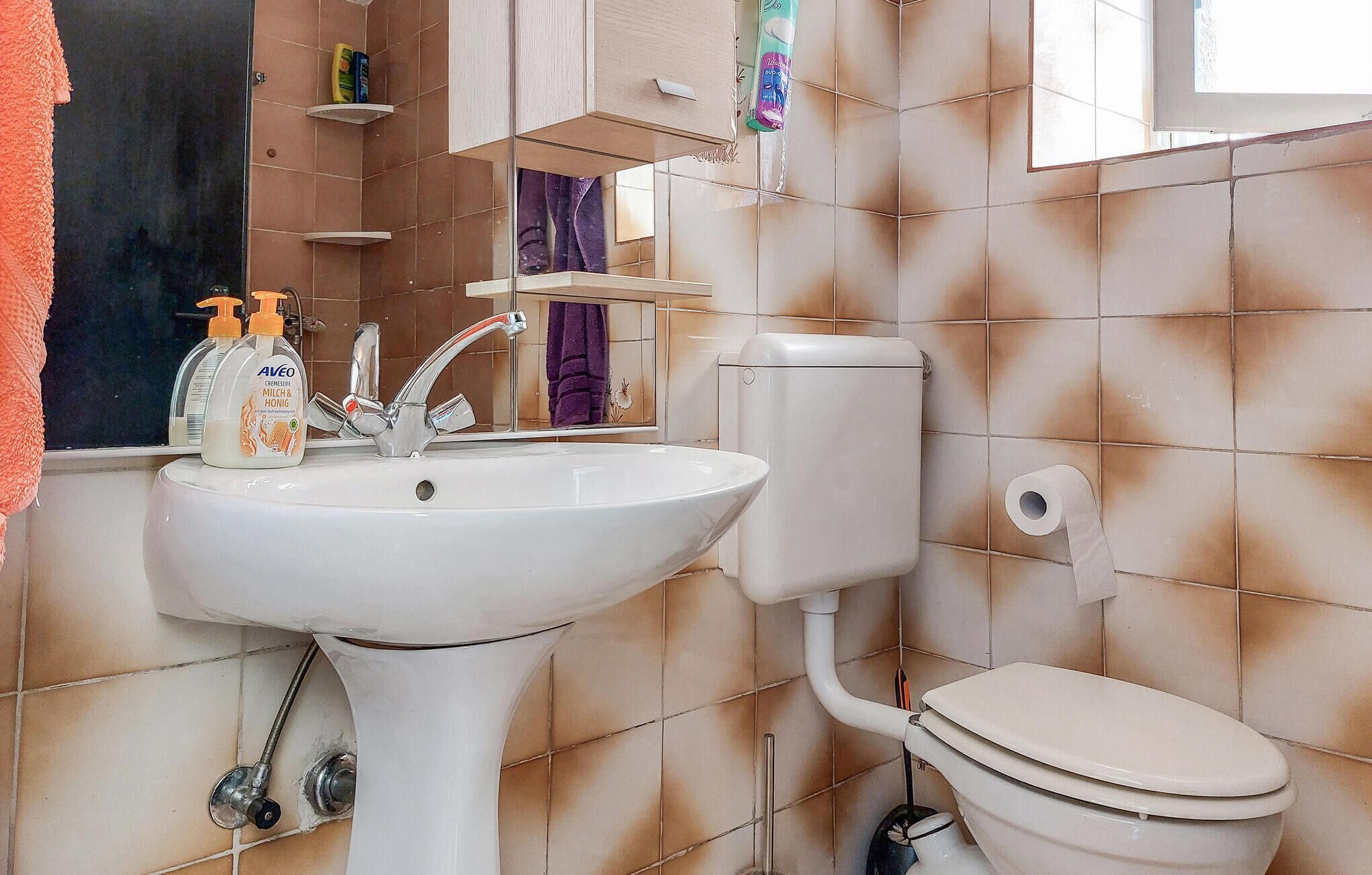 A bathroom with a white sink, a toilet, and a mirror. There are toiletries on the sink and a towel hanging on the wall.