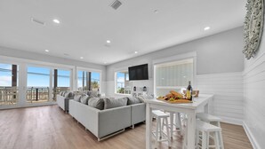 Amplia sala de estar con un sofá gris, una mesa de comedor con una bandeja de frutas y un televisor montado en la pared.