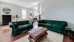 Un salon spacieux avec deux canapés en velours vert, une table basse en bois et un coin repas à l’arrière-plan.