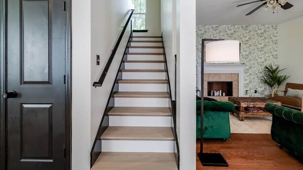 Un escalier avec des marches en bois et des rampes noires, menant à un espace de vie comprenant un canapé vert, une cheminée et un ventilateur de plafond.