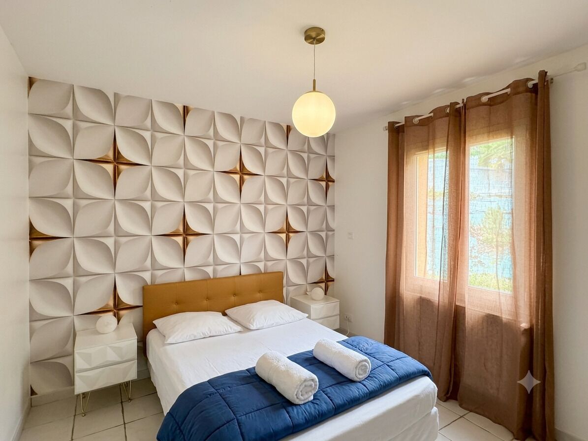 Ein Schlafzimmer mit einem Kopfende mit geometrischem Muster, einem Bett mit blauer Bettdecke, zwei weißen Nachttischen und einer hängenden Leuchte.