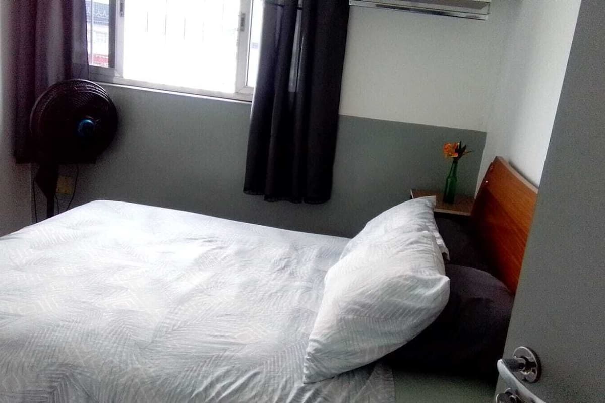 A hotel room with a bed, a nightstand with a vase of flowers, a window with curtains, and a wall-mounted heater.