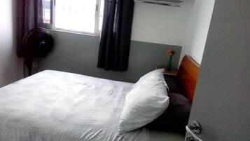 Quarto de hotel com uma cama, mesa de cabeceira com um vaso de flores, janela com cortinas e aquecedor fixado na parede.