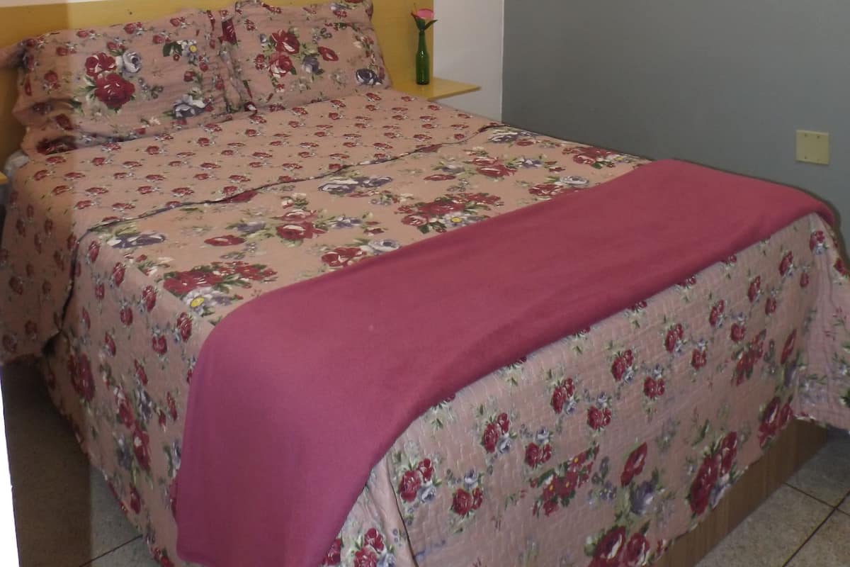 A bedroom with a floral bedspread, a pink blanket, a bedside table with a vase, and a shelf with a rolled-up towel.