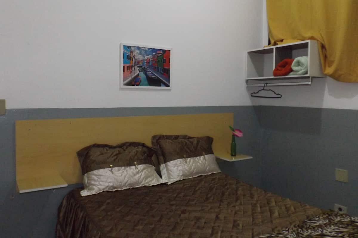 A bedroom with a bed, a painting, a shelf with clothes, and a curtain.