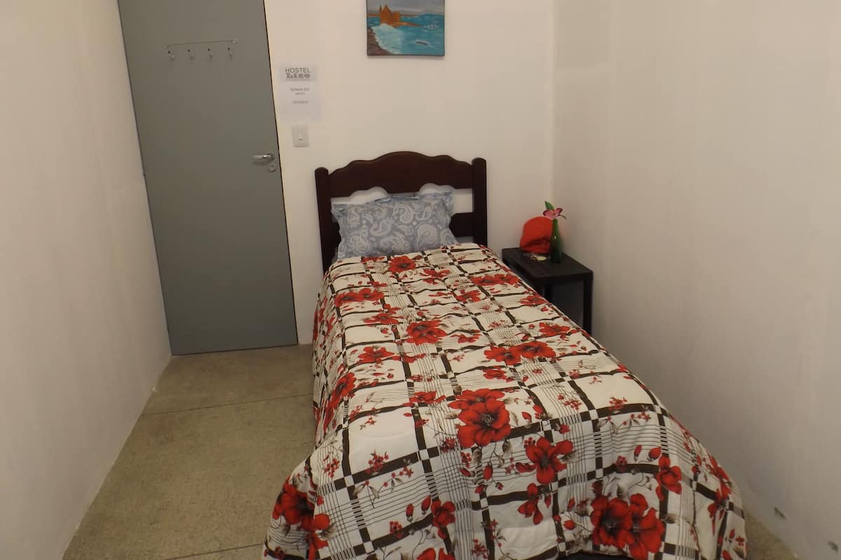 A single bed with a floral bedspread, a nightstand with a vase, a painting on the wall, and a grey door.