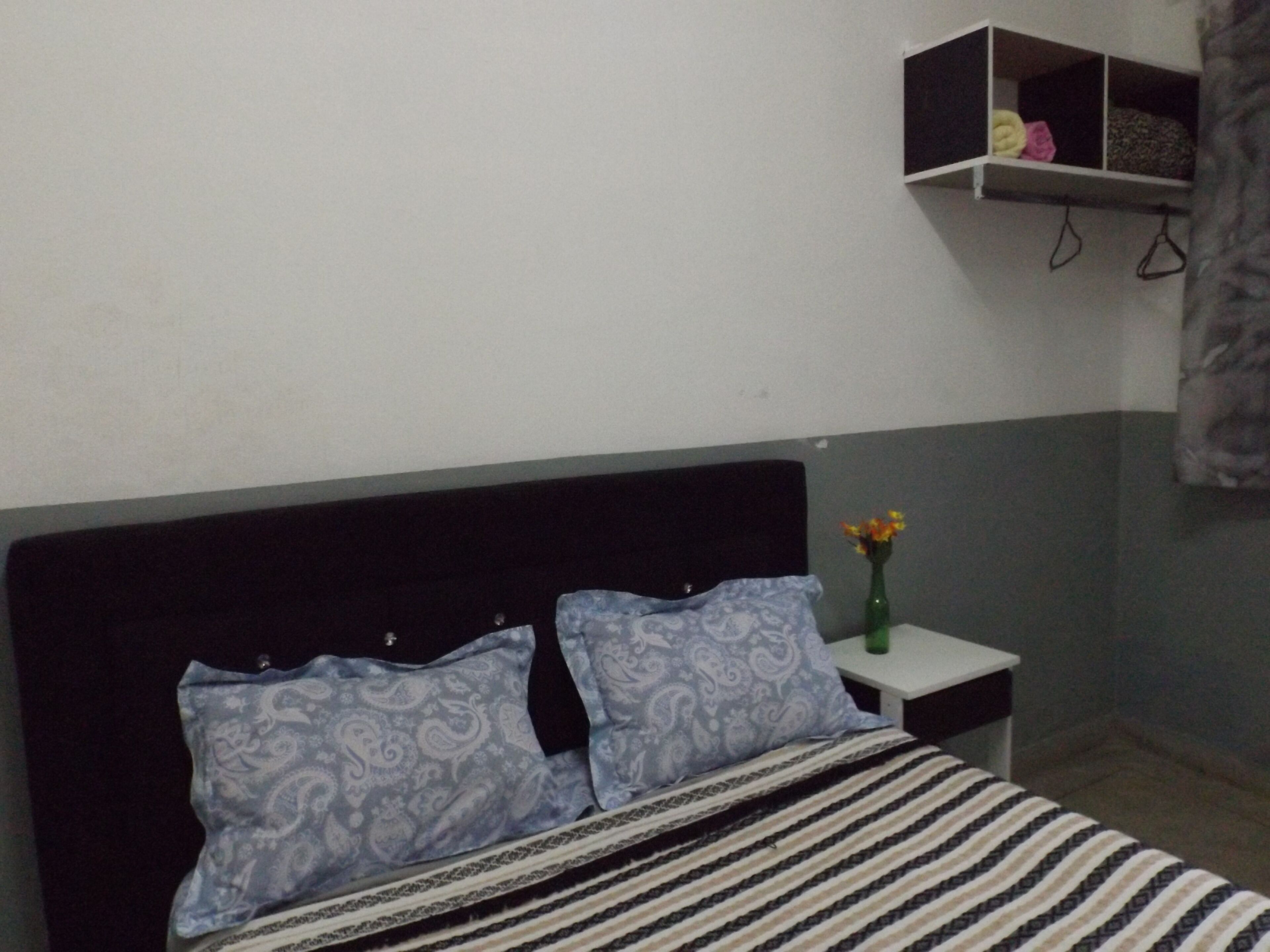Quarto com uma cama, duas almofadas estampadas, uma mesinha de cabeceira com um vaso de flores e uma prateleira sobre a cama.