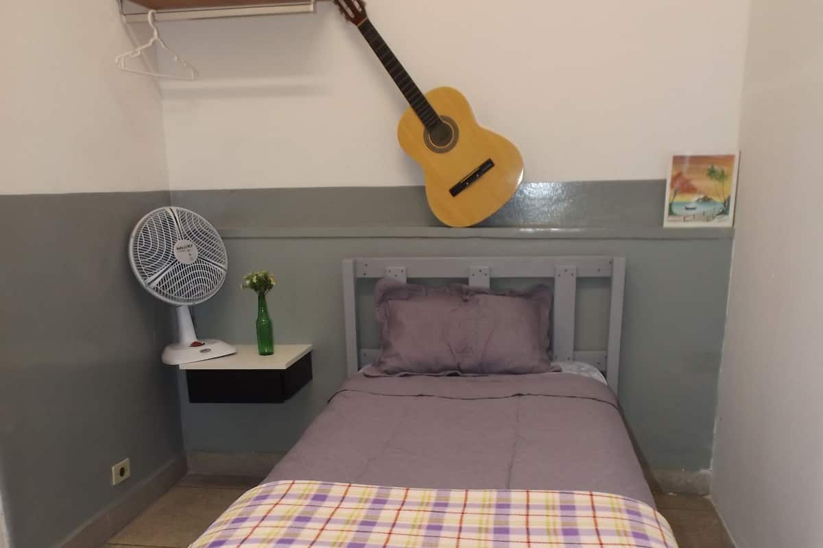 A bedroom with a bed, a guitar, a fan, and a shelf with a towel.