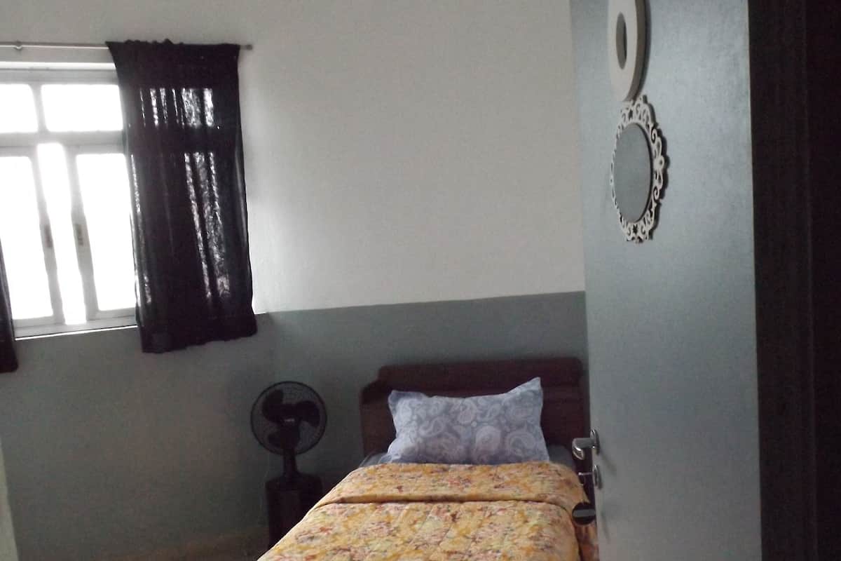 A single bed with a floral bedspread in a room with a window, a wall clock, and a fan.