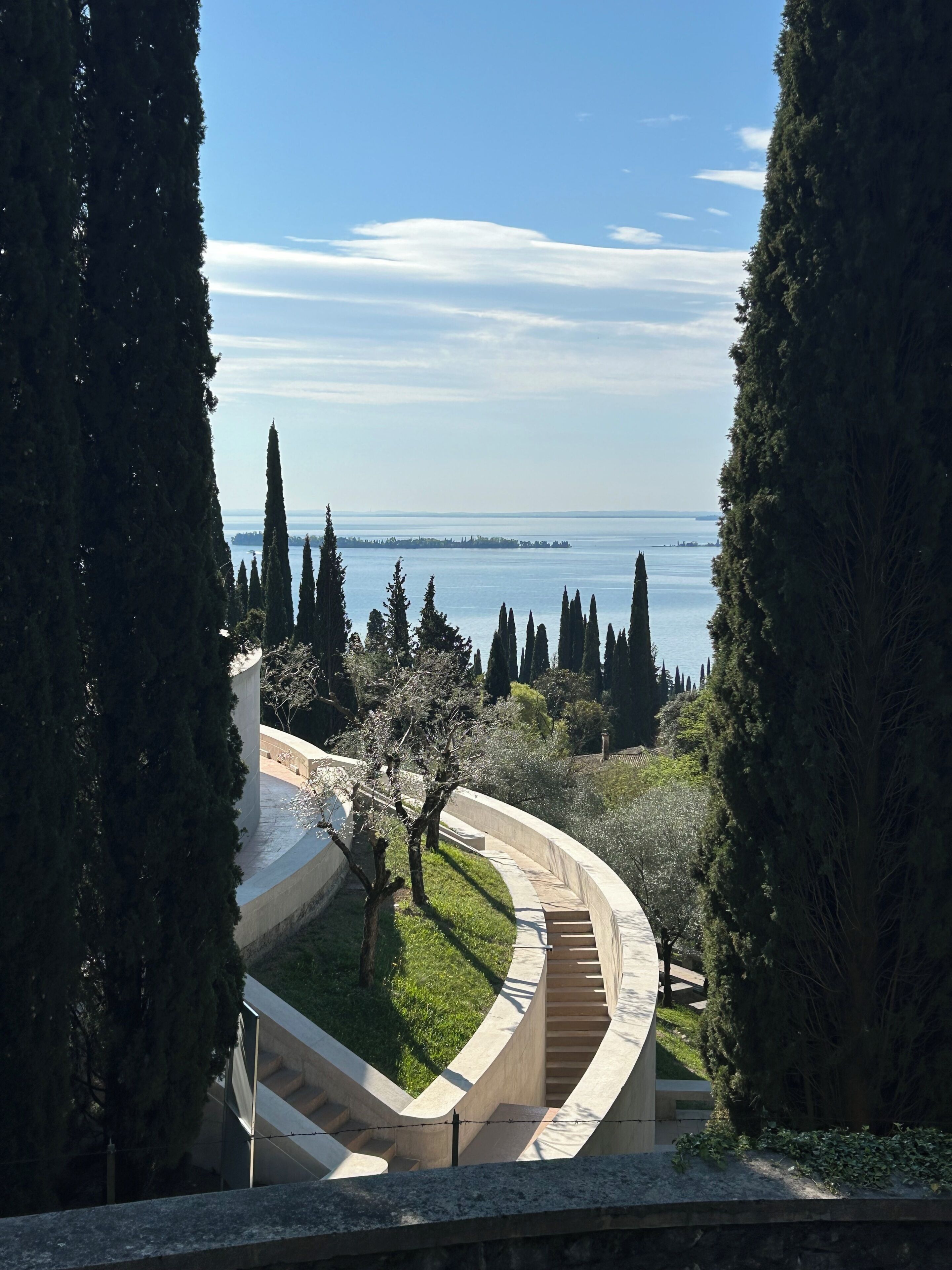 A curved pathway leading to a body of water, surrounded by tall cypress trees and a clear sky.