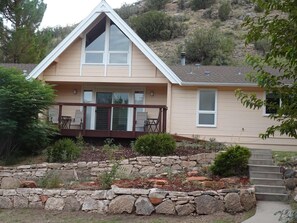 Une maison avec un mur de soutènement en pierre, une véranda couverte avec des chaises et un escalier menant à la porte d’entrée.
