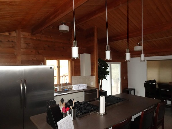 Une cuisine moderne dotée d’un grand îlot central, d’appareils électroménagers en acier inoxydable, de luminaires suspendus et de poutres en bois au plafond.