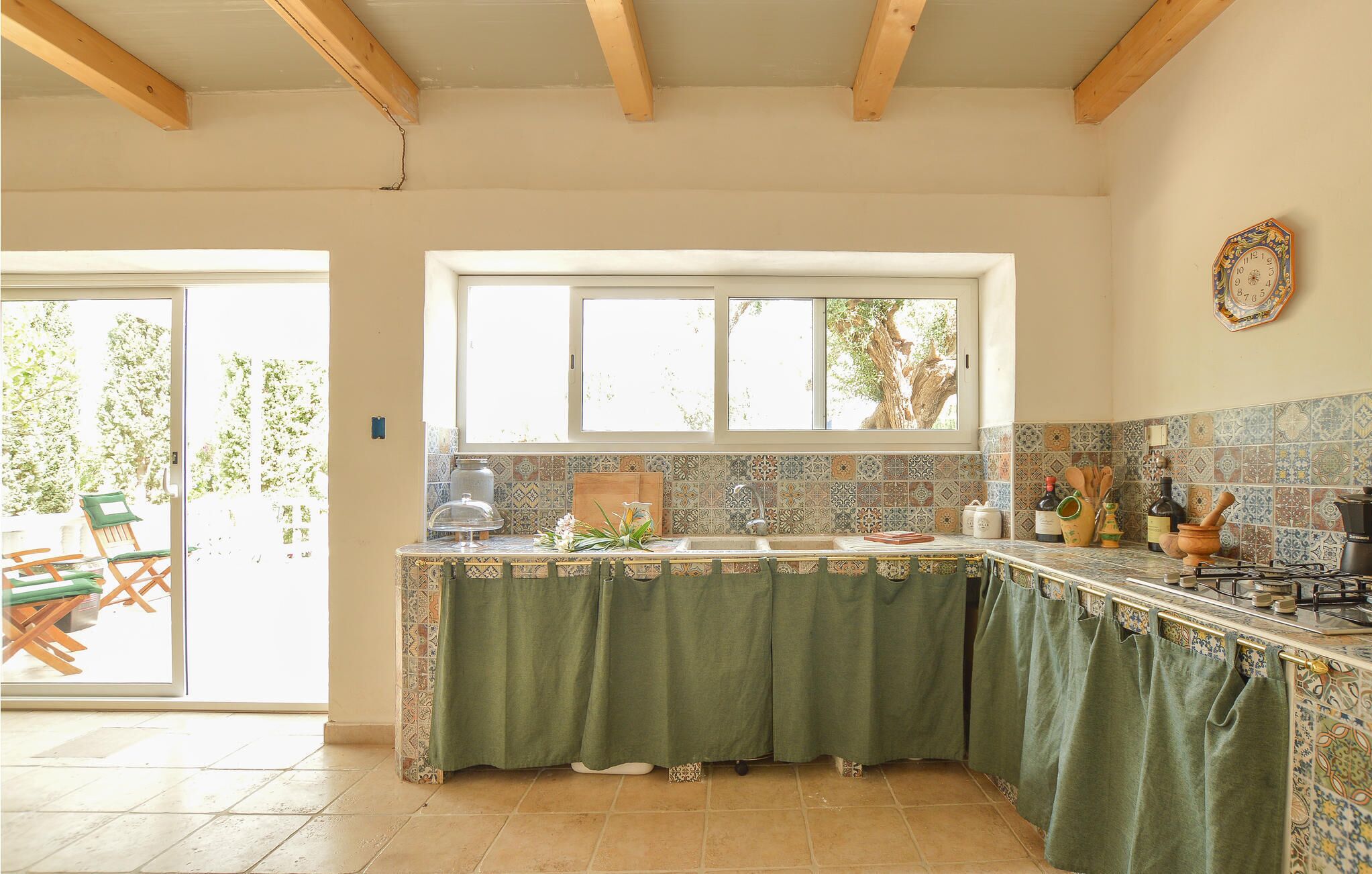 A kitchen with a green curtain, tiled backsplash, and a window overlooking greenery.